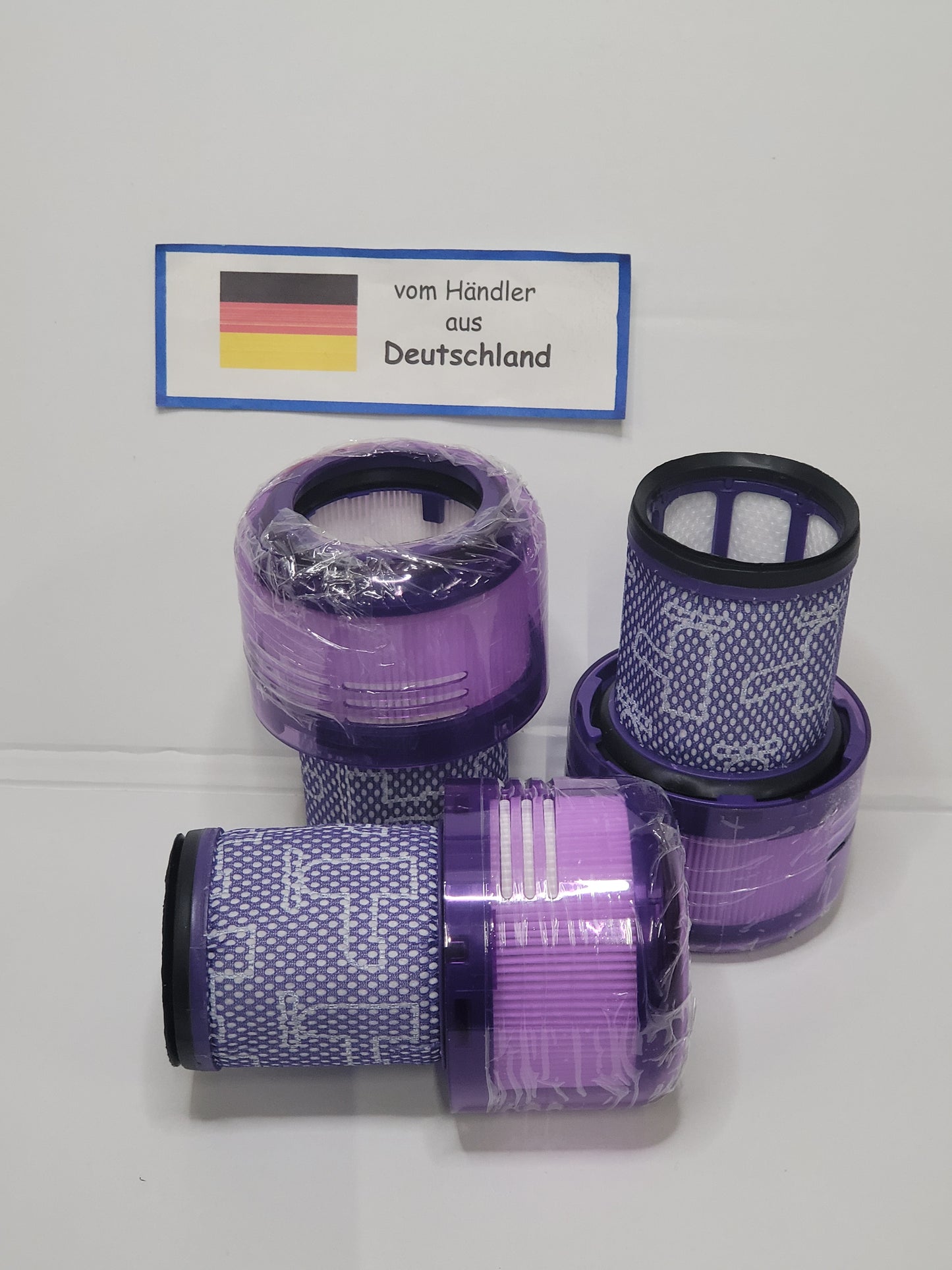 Fünf lilafarbene und weiße zylindrische Staubsaugerfilter, einige davon in Plastik eingewickelt, sind mit einer deutschen Flagge und einem Schild versehen, das darauf hinweist, dass sie von ALMEc Supplies stammen - ideal als Behälter + Filter Dyson V12 Detect slim kompatibel.