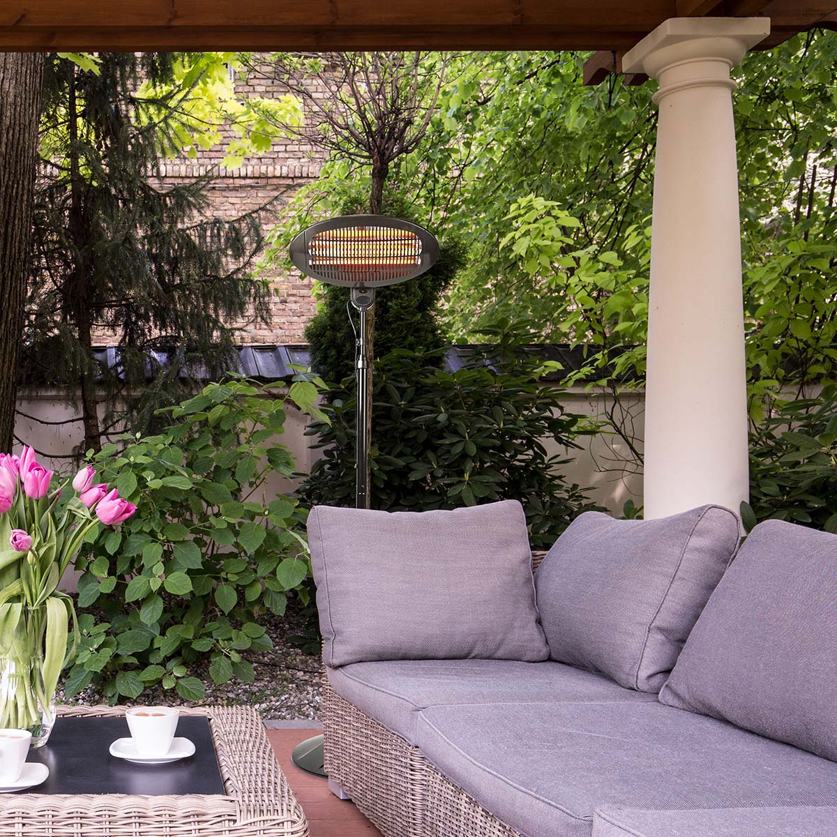 Eine gemütliche Außenterrasse mit einem grauen, gepolsterten Sofa unter einer Pergola aus Holz. Ein stehendes, immersaugfähiges Nedis-Terrassenheizgerät, ausgestattet mit 3 Wärmeeinstellungen und Stolperschutz, steht neben dem Sofa inmitten üppigen Grüns.