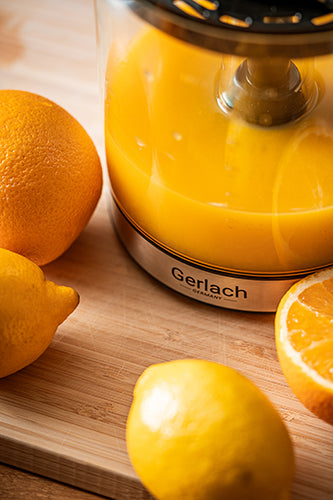 Eine Nahaufnahme der elektrischen Zitruspresse der Adler Europe GmbH (Entsafter, Zitronenpresse, Saftpresse), gefüllt mit frischem Saft, umgeben von ganzen Zitronen und einer halbierten Orange auf einem Holzschneidebrett, die die Zubereitung von frischem Fruchtsaft veranschaulicht.