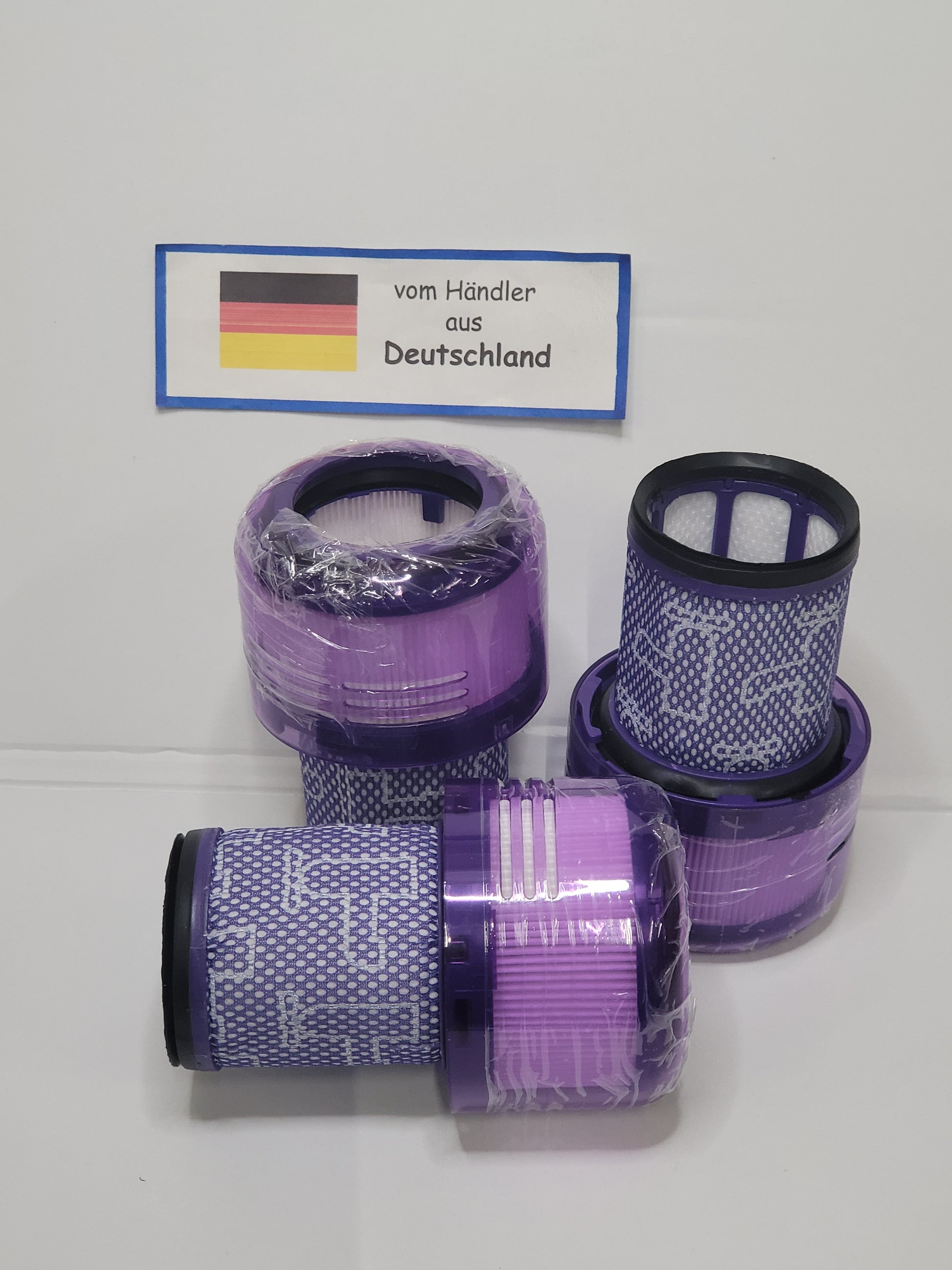 Fünf lilafarbene und weiße zylindrische Staubsaugerfilter, einige davon in Plastik eingewickelt, sind mit einer deutschen Flagge und einem Schild versehen, das darauf hinweist, dass sie von ALMEc Supplies stammen - ideal als Behälter + Filter Dyson V12 Detect slim kompatibel.