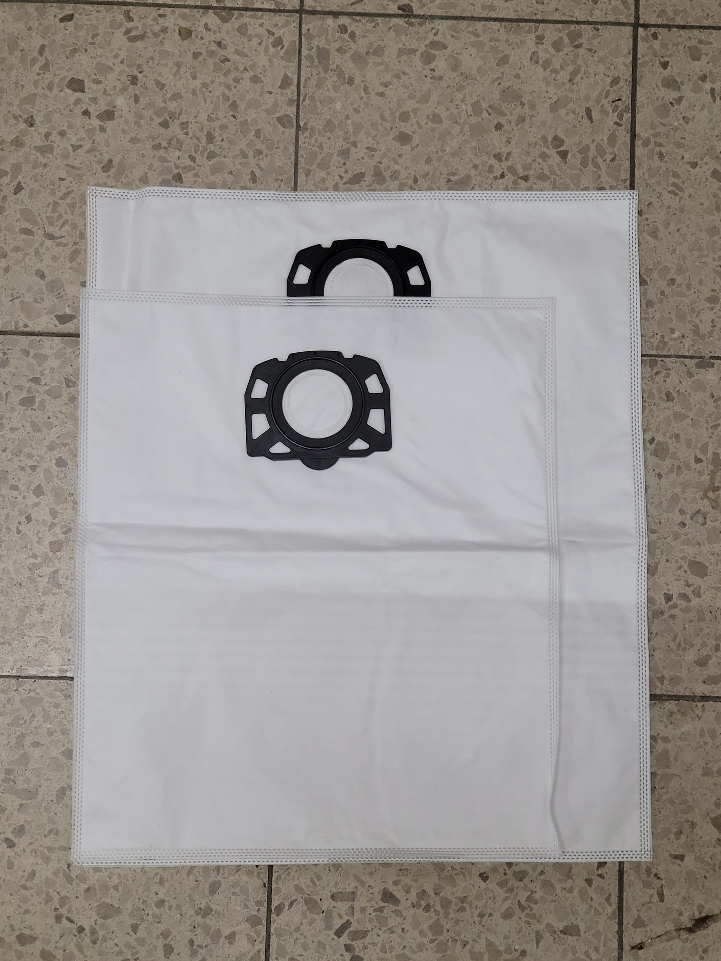 Zwei weiße XXL Staubsaugerbeutel von immer-saugut, aus dem Set "10 Beutel kompatibel mit Kärcher WD6, WD 7", liegen mit schwarzen Plastikmanschetten und sichtbaren Nähten auf einem beige gesprenkelten Fliesenboden, leicht überlappend.