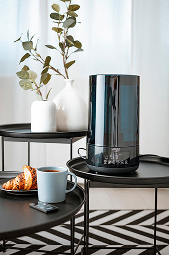 Ein „immer-saugut“-Luftbefeuchter AD 7963 in Schwarz mit 4,3 Litern Fassungsvermögen und Aromatherapie-Funktion steht auf einem runden schwarzen Beistelltisch neben einer weißen Vase, die mit grünen Blättern gefüllt ist. Daneben steht ein weiterer Tisch, auf dem ein blauer Becher, zwei Croissants auf einem Teller und ein Smartphone liegen. Im Hintergrund sind weiße Vorhänge und geometrische Teppichmuster zu sehen, während die Fernbedienung des Luftbefeuchters bequem in Reichweite liegt.