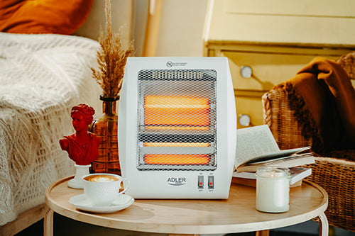 Auf einem runden Holztisch stehen ein Halogen-Heizstrahler vom Typ MS 7709 der Firma Adler Europe GmbH, ein Cappuccino, eine brennende Kerze, ein aufgeschlagenes Buch und eine rote Dekobüste. Gemütliche Decken und Kissen im Hintergrund sorgen für eine warme, einladende Atmosphäre.