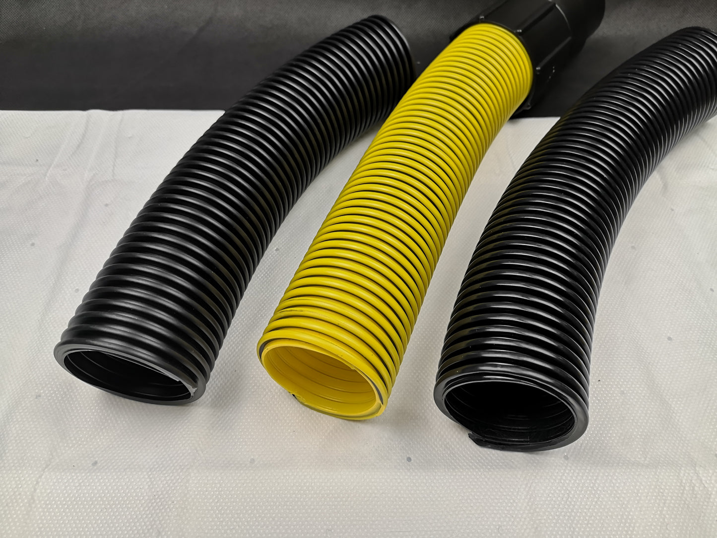 Auf einer weißen Fläche sind drei zylindrische, flexible Wellschläuche abgebildet. Der mittlere Schlauch ist der gelbe, geriffelte immersaugende Saugschlauch Meterware 51 mm, während der linke und rechte Schlauch schwarz sind.