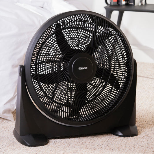 Ein schwarzer runder Mesko - Bodenventilator - 3 Leistungsstufen, XXL Rotor 50 cm, Neigungsfunktion von immer-saugut mit Maschengitter und fünf Flügeln steht auf einem beigen Teppichboden, neben einem Bett mit weißer Bettwäsche. Sein leistungsstarker Moto