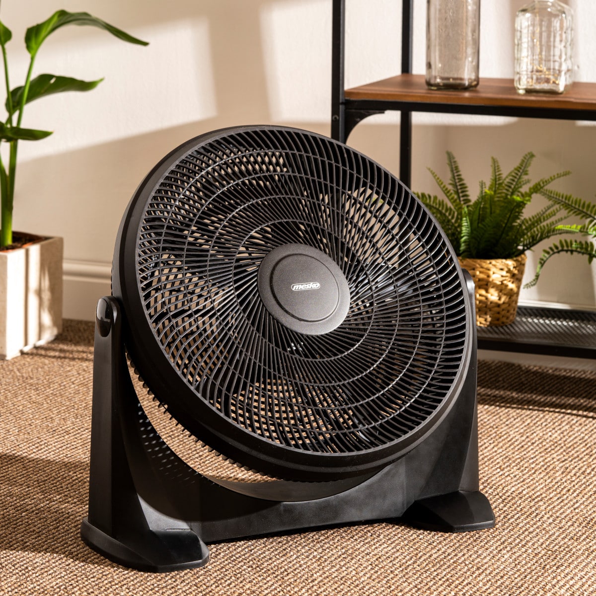 Ein großer schwarzer **immer-saugut Mesko - Bodenventilator - 3 Leistungsstufen, XXL Rotor 50 cm, Neigungsfunktion** mit einstellbarem Neigungskopf steht auf einem beigen Teppich in einem gut beleuchteten Raum. Hinter dem Ventilator steht links eine Topfp