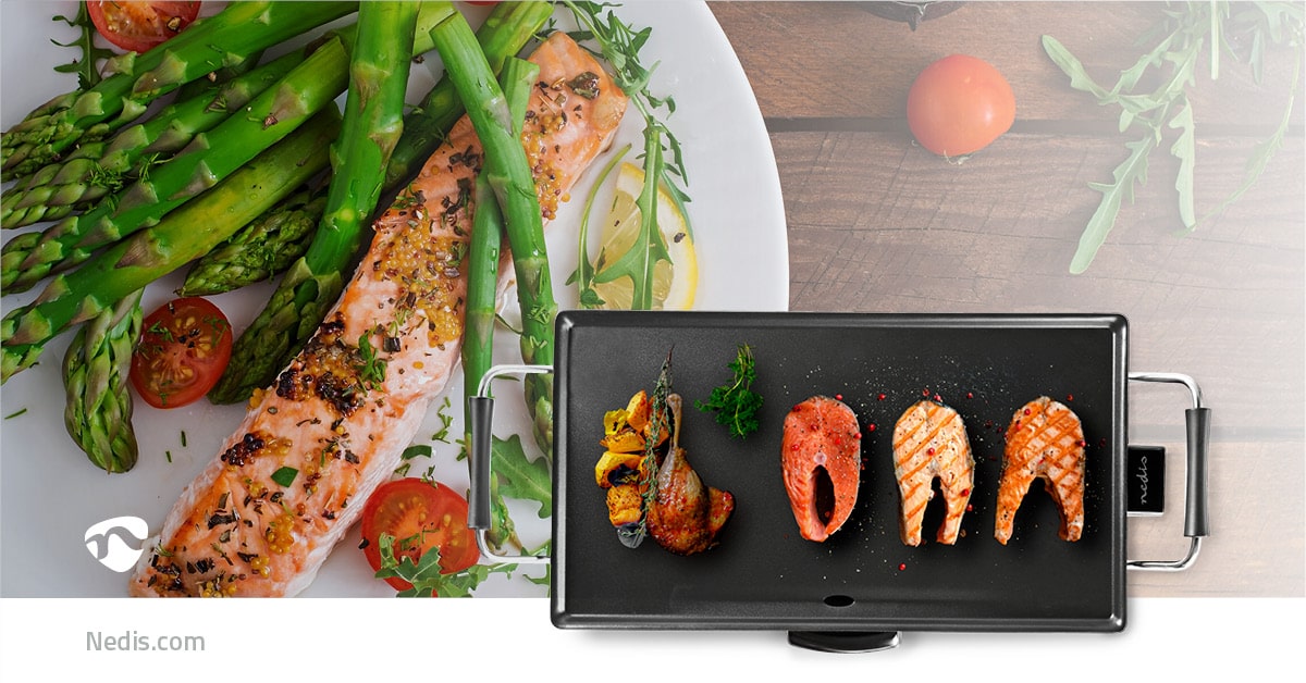 Auf einem Holztisch steht ein Teller mit gekochtem Lachs, Spargel und Tomaten. Rechts daneben liegt ein immer-saugut Teppanyaki-Tischgrill mit drei Lachsstücken, einer Orangenscheibe und einigen Kräutern, die in die Antihaft-Beschichtung eingelegt sind. I