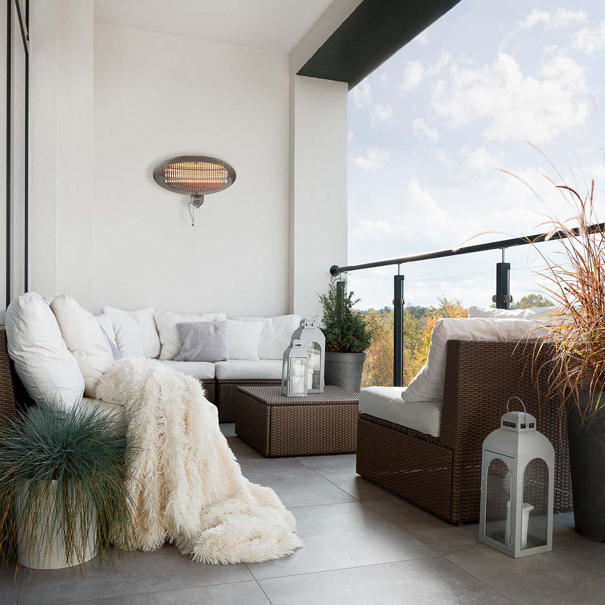 Ein gemütlicher Balkon mit weißen Korbmöbeln und Kissen in L-Form. Über dem Sofa hängt eine flauschige cremefarbene Decke. In der Mitte steht ein Couchtisch mit einer Laterne. Hohe Topfpflanzen und ein an der Wand montierter Nedis-Terrassenheizer von imme