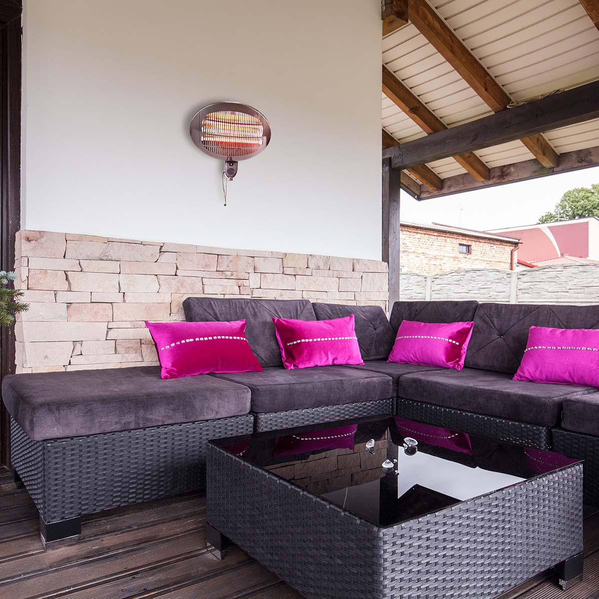 Ein gemütlicher Sitzbereich im Freien verfügt über ein dunkles Korbsofa mit Kissen in leuchtendem Lila und Magenta. Vor dem Sofa steht ein quadratischer Korb-Couchtisch mit Glasplatte. Der Raum ist teilweise umschlossen, mit einer Akzentwand aus Stein und