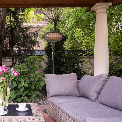 Eine gemütliche Außenterrasse mit einem grauen, gepolsterten Sofa unter einer Pergola aus Holz. Ein stehendes, immersaugfähiges Nedis-Terrassenheizgerät, ausgestattet mit 3 Wärmeeinstellungen und Stolperschutz, steht neben dem Sofa inmitten üppigen Grüns.