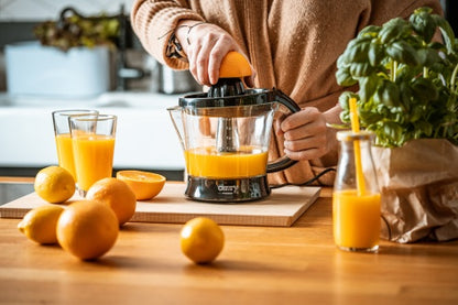 Eine Person verwendet eine 40-W-Elektrische Zitruspresse CR 4008 der Firma Adler Europe GmbH, um frisch gepressten Saft aus frischen Orangen herzustellen. Auf dem Holztisch liegen ganze Orangen, eine aufgeschnittene Orange, zwei Gläser und eine Saftflasche mit einem gelben Strohhalm, daneben steht eine Papiertüte mit Basilikum.
