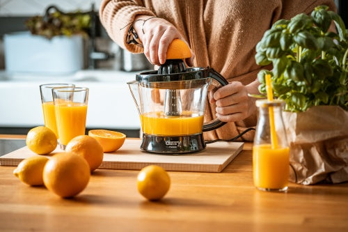 Eine Person verwendet eine 40-W-Elektrische Zitruspresse CR 4008 der Firma Adler Europe GmbH, um frisch gepressten Saft aus frischen Orangen herzustellen. Auf dem Holztisch liegen ganze Orangen, eine aufgeschnittene Orange, zwei Gläser und eine Saftflasche mit einem gelben Strohhalm, daneben steht eine Papiertüte mit Basilikum.