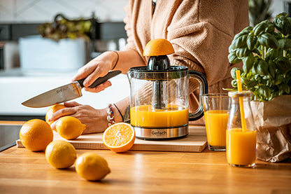 Eine Person schneidet eine Orange auf einem Schneidebrett neben einer elektrischen Zitruspresse der Adler Europe GmbH, die mit frischem Saft gefüllt ist. Daneben liegen ganze und halbierte Orangen. Ein Glas Saft mit einem gelben Strohhalm und eine Basilikumpflanze stehen auf der Holztheke und schaffen eine helle und gemütliche Atmosphäre.